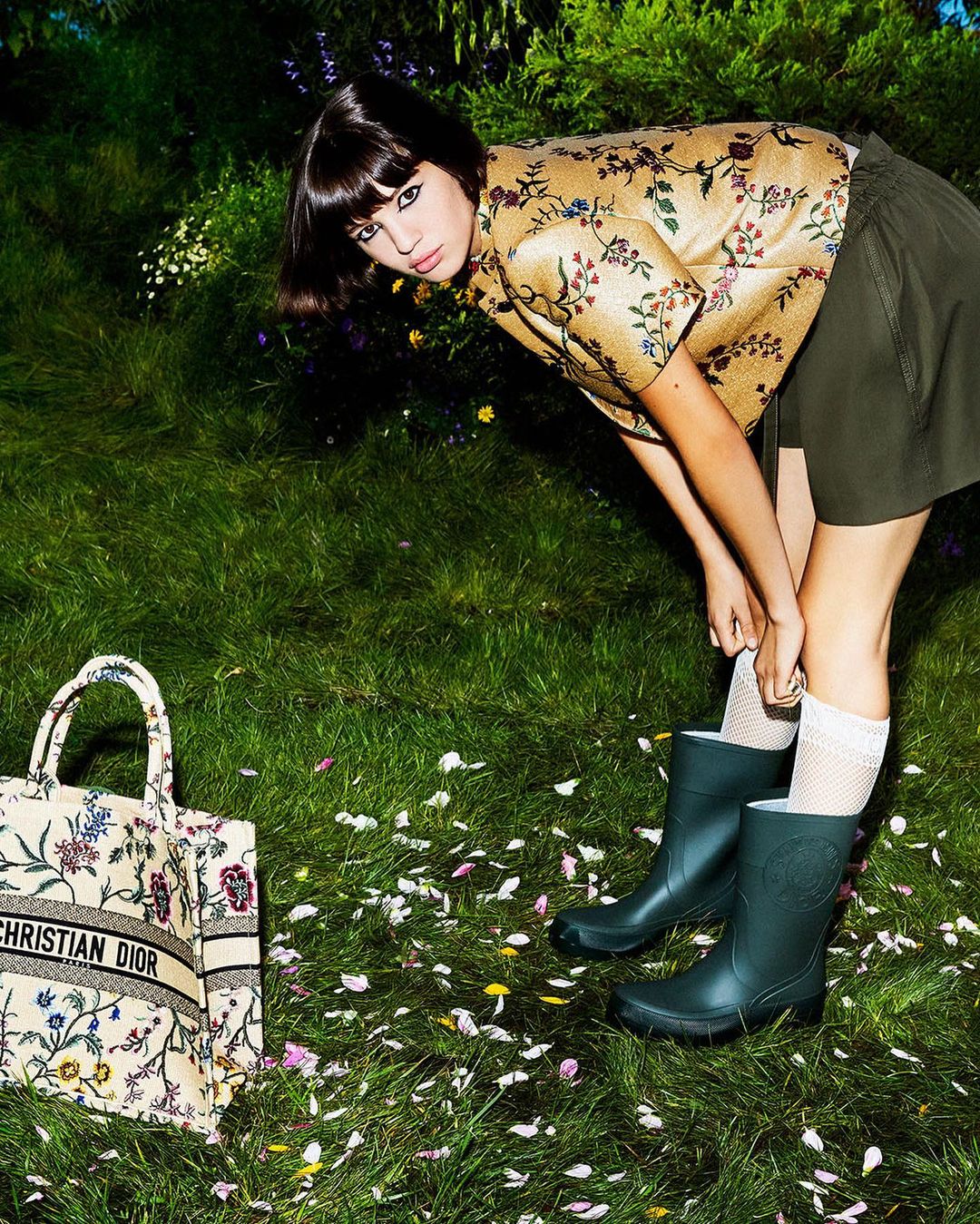Woman poses in a floral top and skirt, adjusting her rain boot on a grass lawn with a Christian Dior tote bag nearby.