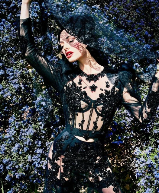 Woman in elaborate black lace gown and veil poses among purple flowers, creating a high fashion, editorial, and ethereal outdoor scene.