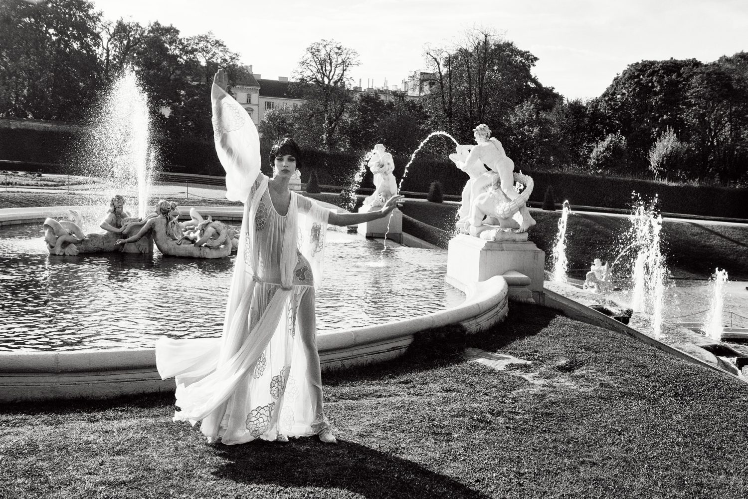 Elegant woman in flowing white dress poses near ornate fountain in a black and white photograph on manicured grounds of a European estate.