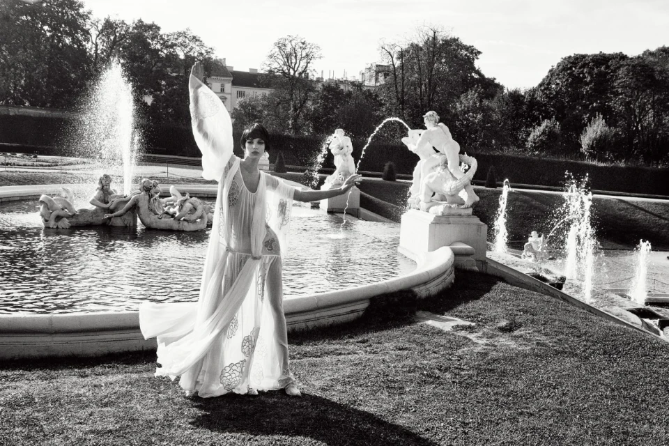 Elegant woman in flowing white dress poses near ornate fountain in a black and white photograph on manicured grounds of a European estate.