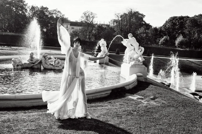 Elegant woman in flowing white dress poses near ornate fountain in a black and white photograph on manicured grounds of a European estate.
