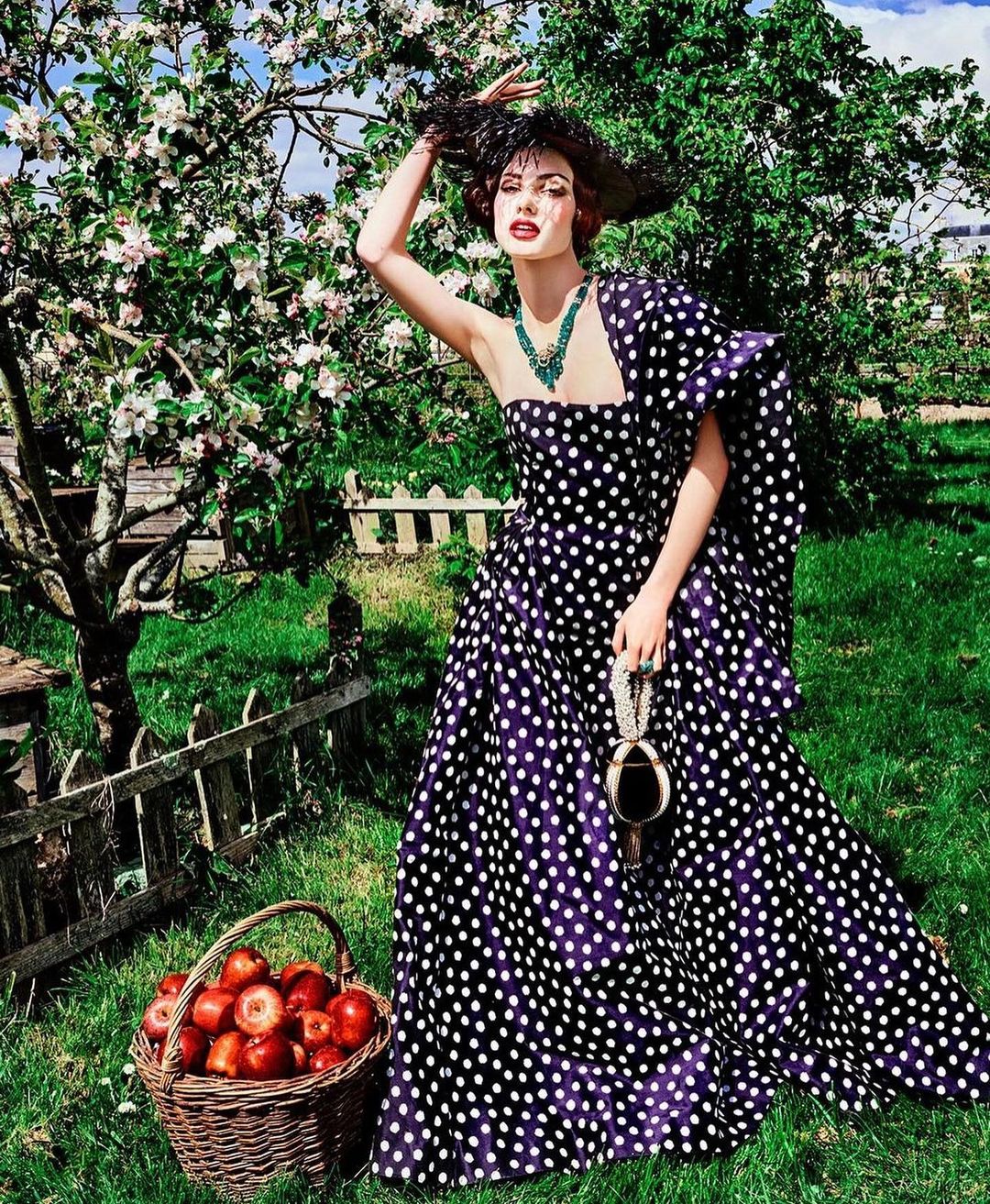 A woman wearing a polka dot dress and a feathered hat is standing in a garden next to an apple tree and a basket of red apples.