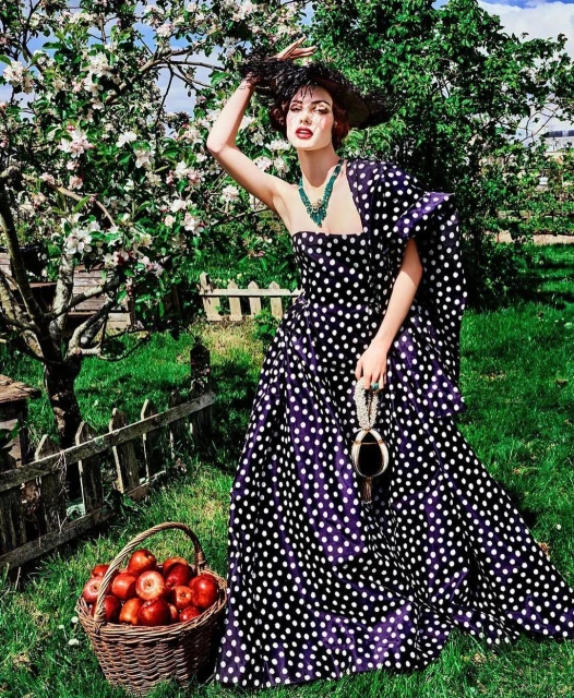 A woman wearing a polka dot dress and a feathered hat is standing in a garden next to an apple tree and a basket of red apples.
