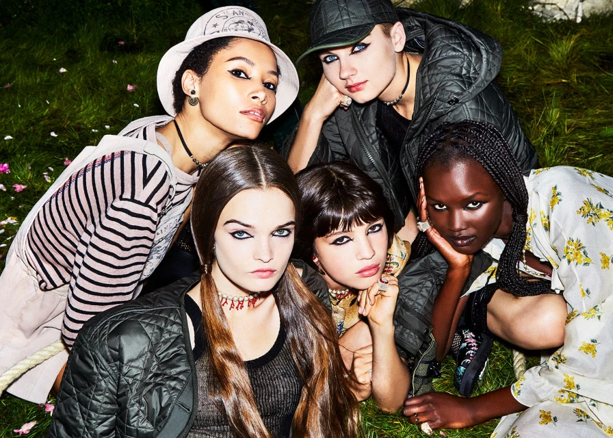 Group of five diverse young women posing outdoors with intense makeup, wearing fashionable clothing and accessories, and green background.