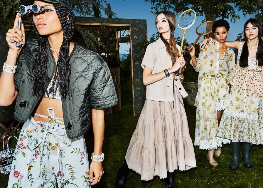 A group of five women in an outdoor setting, wearing dresses and jackets with floral and quilted patterns, and holding binoculars.