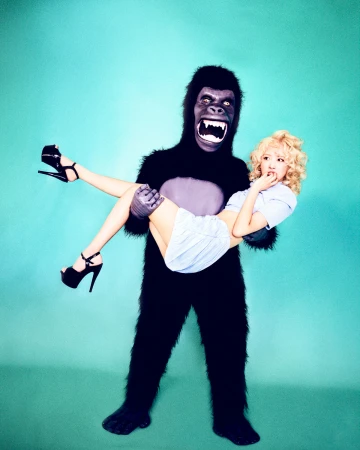Woman in a light blue dress being held in the arms of a person wearing a gorilla costume against a blue gradient background.
