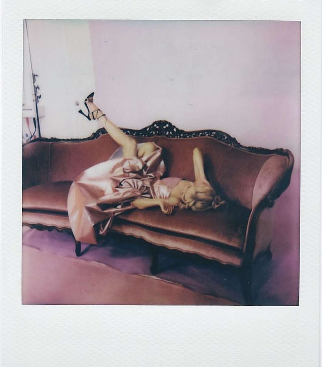 Adult female in a peach dress and black heels poses on a plush velvet couch inside a room, captured with a Polaroid effect.