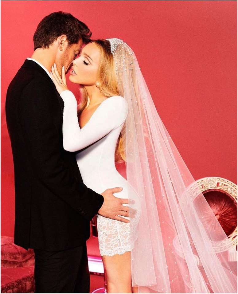 Bride in white dress and long veil embraces groom in suit against red backdrop, photographed in a studio setting with bright, clean lighting.