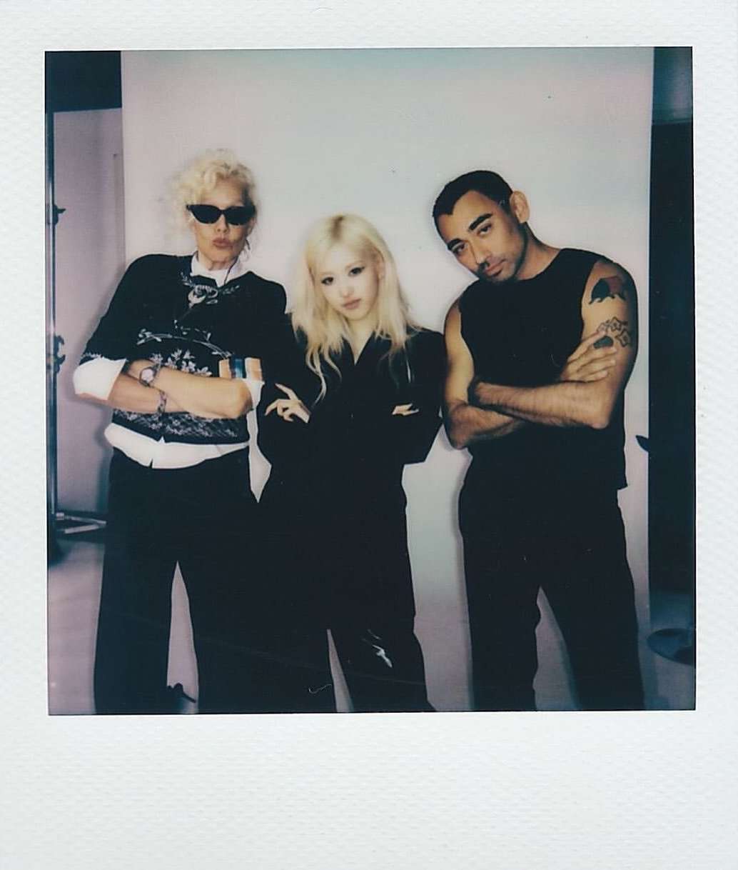 Three adults pose indoors with arms crossed, wearing casual attire against a plain backdrop, captured in a photo studio setting.