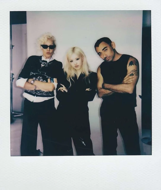 Three adults pose indoors with arms crossed, wearing casual attire against a plain backdrop, captured in a photo studio setting.