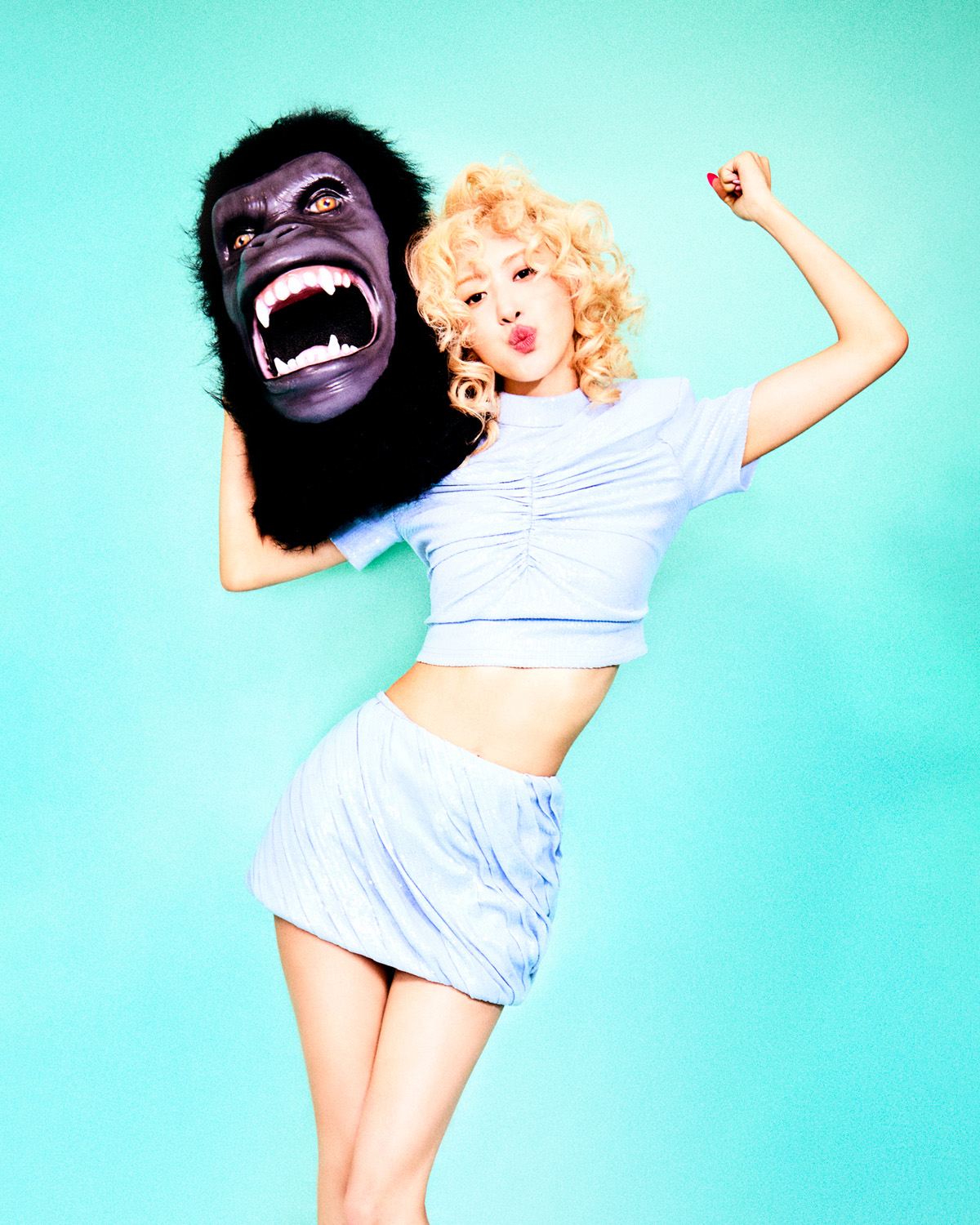 Blonde woman in a light blue outfit poses with a large gorilla head against an aqua background, raising her fist in a fun, playful shot.