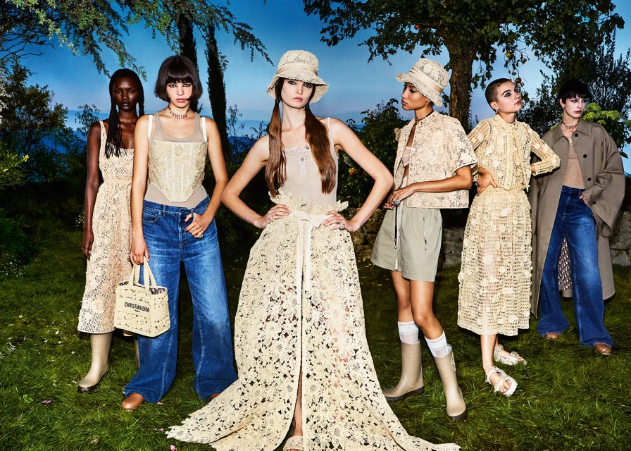 Multiple women in matching earth-tone lace clothing pose in an outdoor, naturalistic setting with a bright blue sky, trees, and greenery.