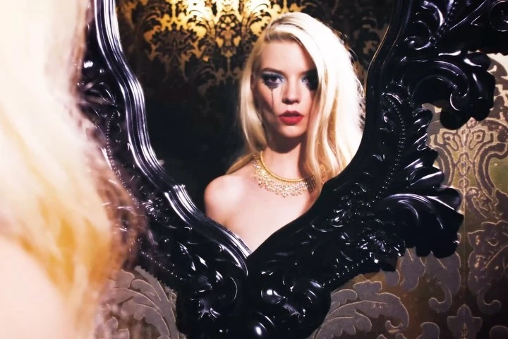Woman with platinum blonde hair, red lipstick, and black teardrop makeup stands reflecting in an ornate black mirror against patterned wallpaper.