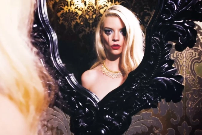 Woman with platinum blonde hair, red lipstick, and black teardrop makeup stands reflecting in an ornate black mirror against patterned wallpaper.