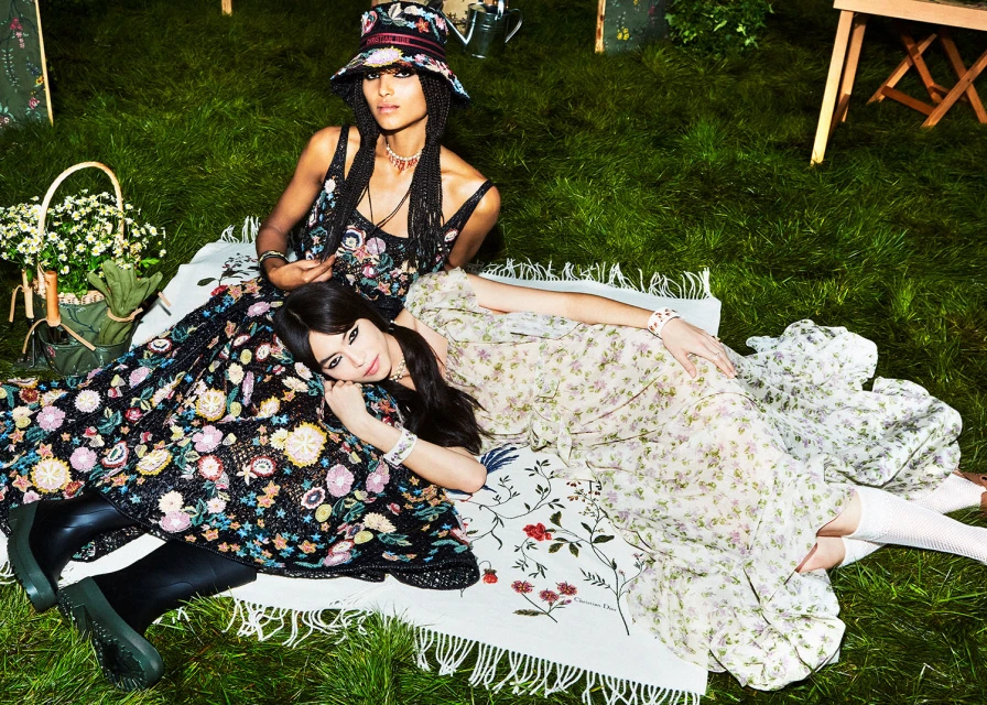 Two women in floral dresses and hats sit on a blanket in a grassy field with white flowers and foliage, fashion editorial.
