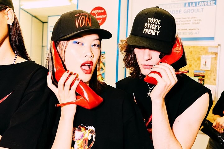 Two young adults with painted faces are holding red stiletto shoes to their ears like telephones indoors, in front of a bulletin board.