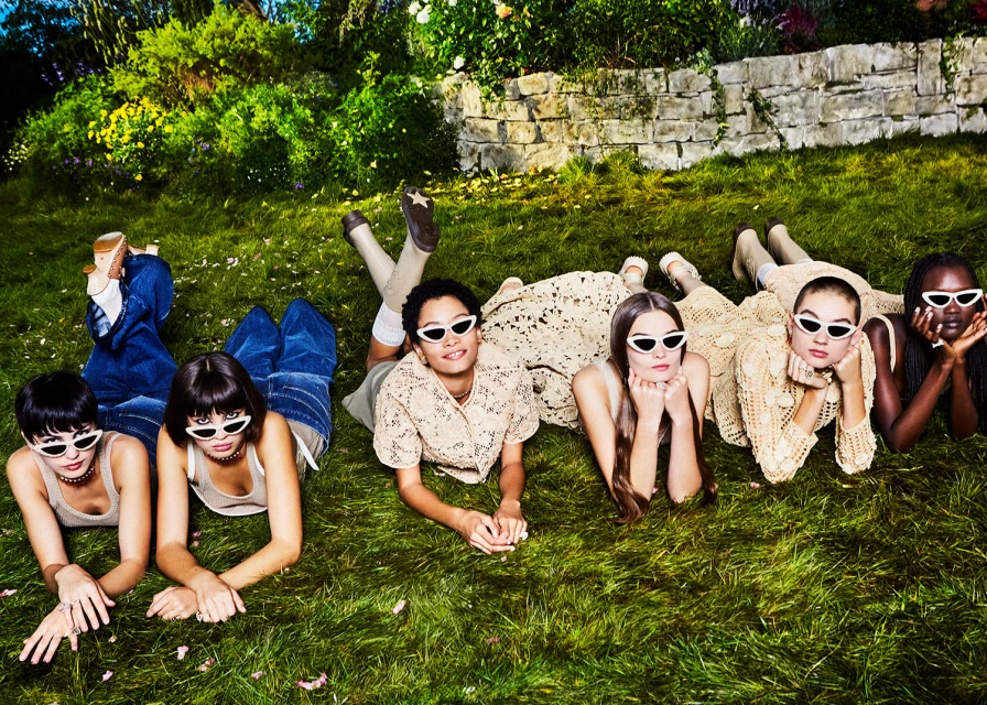 Group of six young adult women in sunglasses lie on grass in neutral-tone outfits in outdoor setting with stonewall and green foliage.