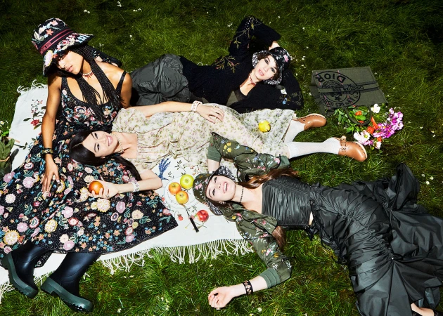 Four fashion models in floral dresses and bucket hats pose on a floral picnic blanket surrounded by greenery and flowers outdoors.