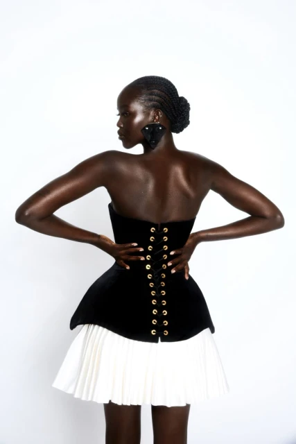 A Black woman poses in a black corset top with gold eyelets and a white pleated skirt against a white background in a studio shot.