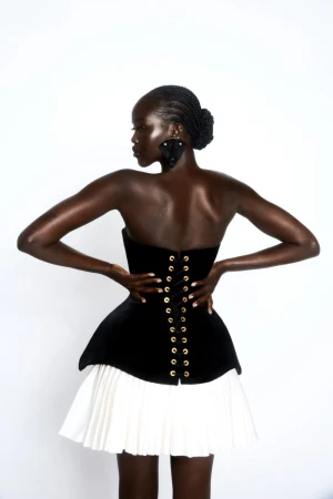 A Black woman poses in a black corset top with gold eyelets and a white pleated skirt against a white background in a studio shot.