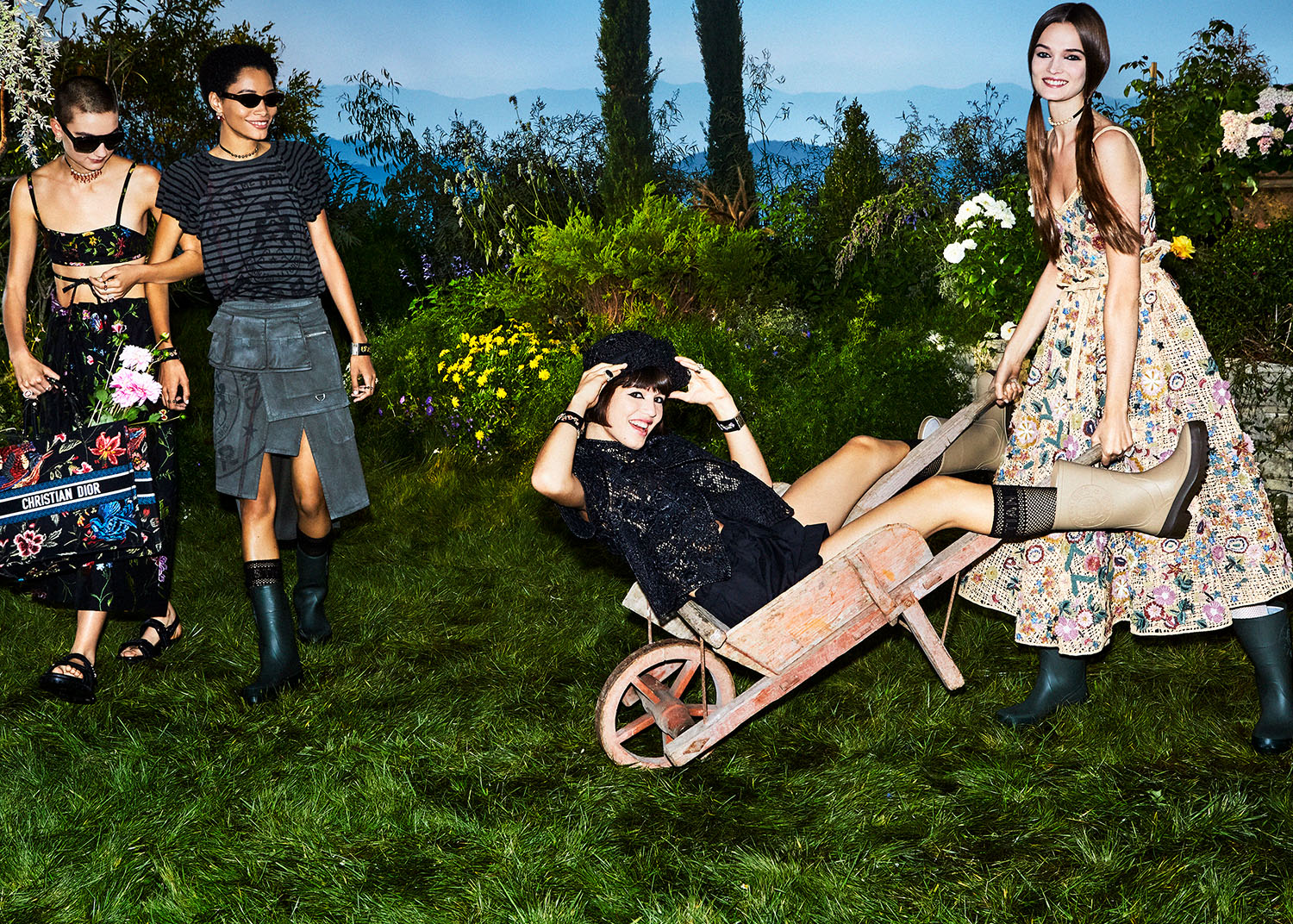 Four women model floral and black fashions in a lush green garden setting with flowers, trees, and a wheelbarrow on a bright day.