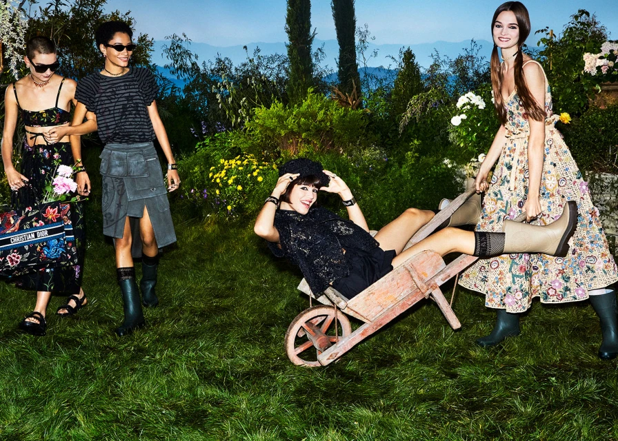 Four women model floral and black fashions in a lush green garden setting with flowers, trees, and a wheelbarrow on a bright day.