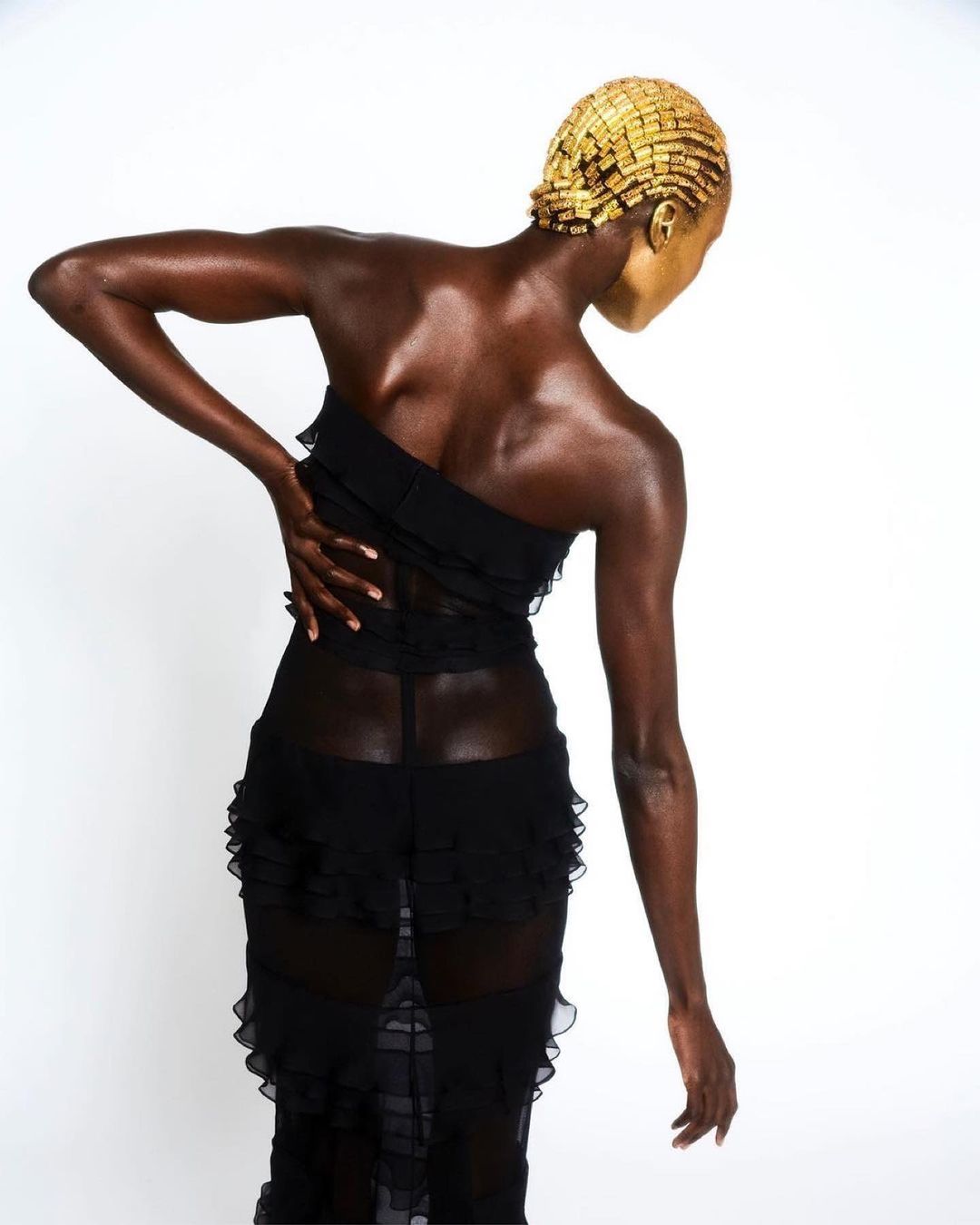 Back view of a woman in a black sheer ruffled dress with golden face and head adornment standing against a white backdrop.
