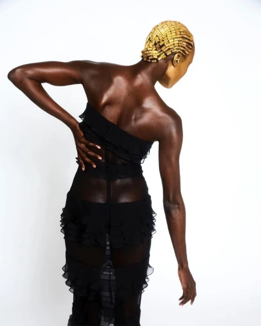 Back view of a woman in a black sheer ruffled dress with golden face and head adornment standing against a white backdrop.