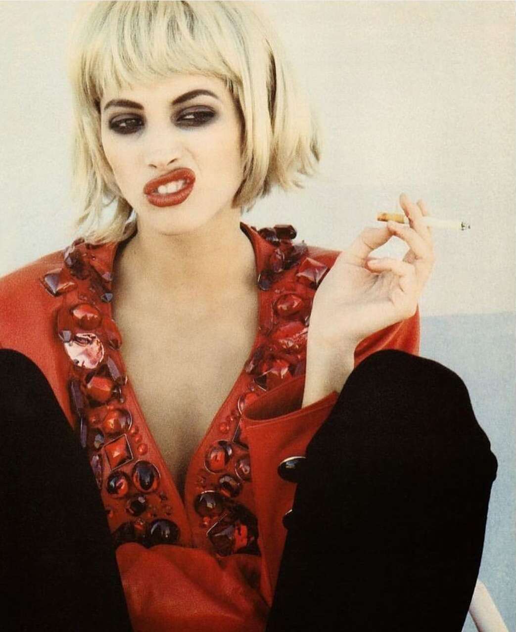 Blonde woman with dark makeup smoking a cigarette and making a face while wearing a red embellished top