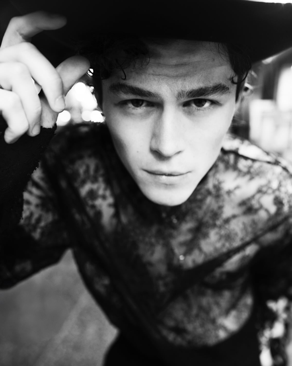 Black and white portrait of a young adult man wearing a lace shirt and wide-brimmed hat looking at the camera in an outdoor setting.