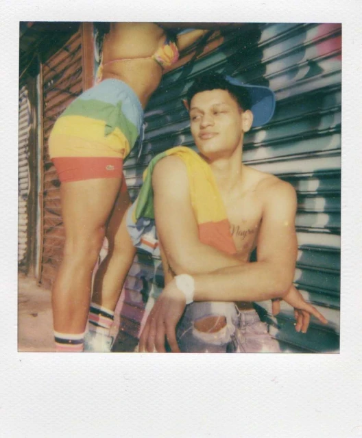 Polaroid photo of a shirtless man in jeans and a hat with a colorful towel, next to a person in a rainbow bikini and shorts outdoors.