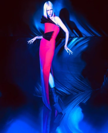 A woman with platinum blonde hair stands in a vibrant red gown with black accents, under a blue spotlight, striking a fashion pose.