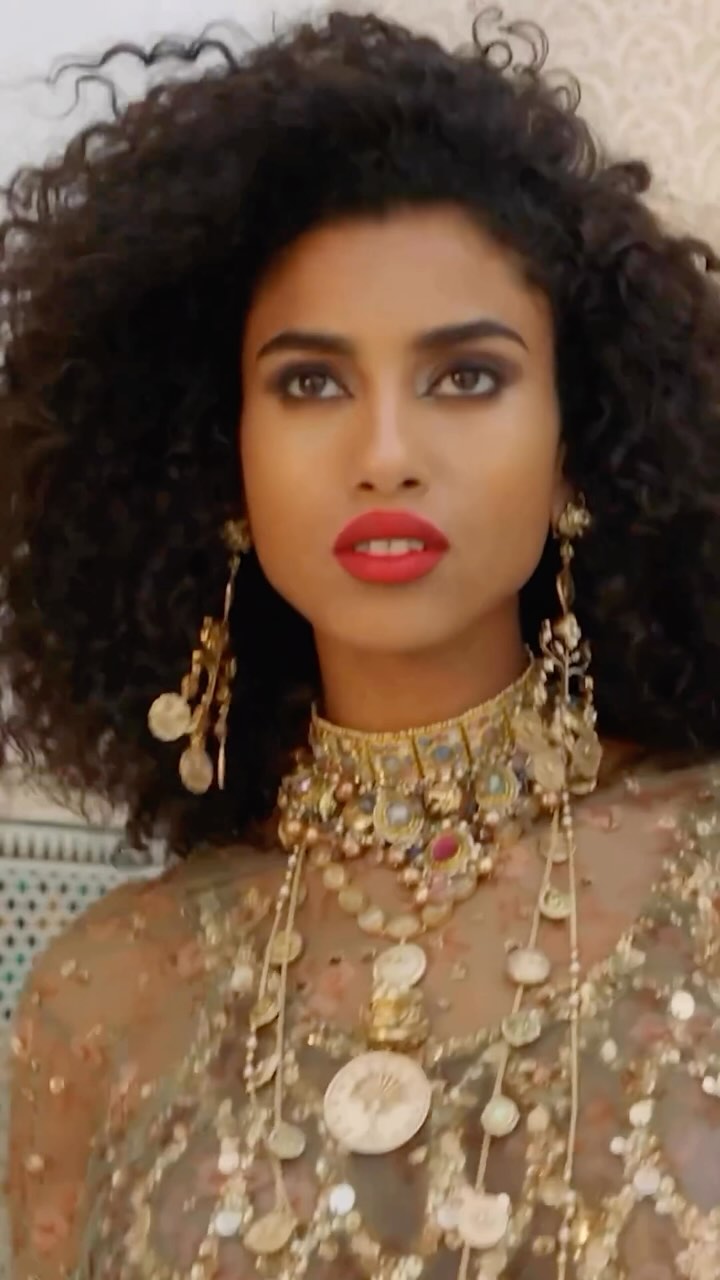 Portrait of an adult female with dark, curly hair wearing a gold choker and necklace with coin details against a patterned backdrop.