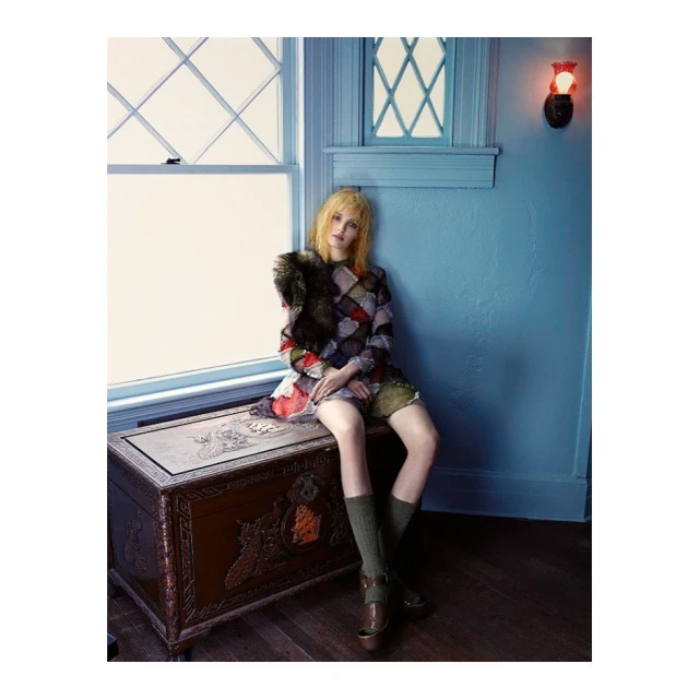 Young woman with blonde hair seated on a decorative trunk, wearing a patchwork dress and knee-high socks, posed near a window.