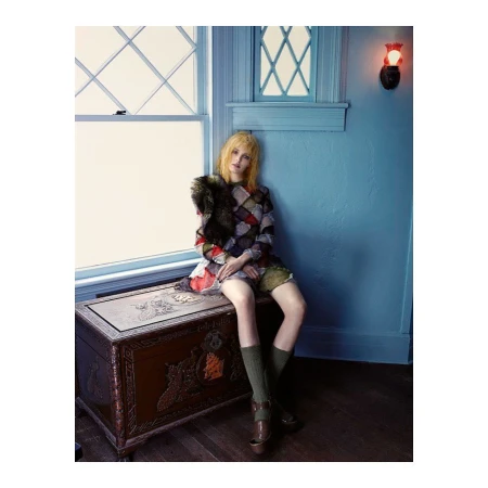 Young woman with blonde hair seated on a decorative trunk, wearing a patchwork dress and knee-high socks, posed near a window.