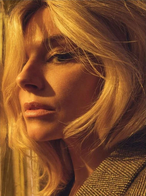 Close-up portrait of a blonde woman with a serious expression. Her golden hair is highlighted in warm lighting, draped over a patterned jacket.