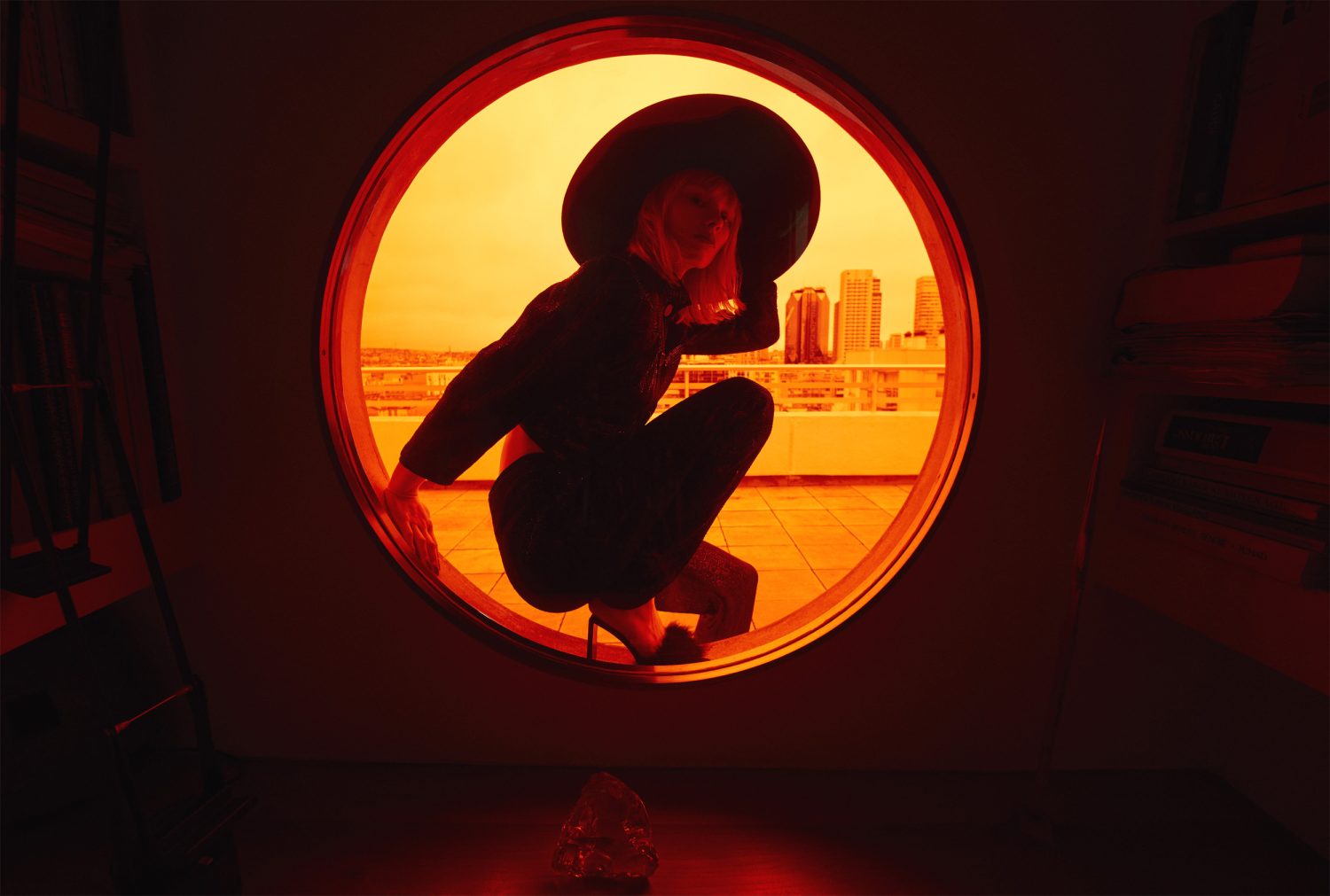 Woman in wide-brim hat poses in circular window on rooftop, city skyline visible in golden light, red color-grading, architectural setting.