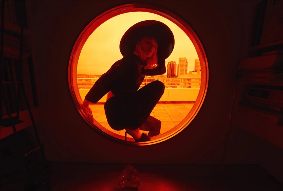 Woman in wide-brim hat poses in circular window on rooftop, city skyline visible in golden light, red color-grading, architectural setting.