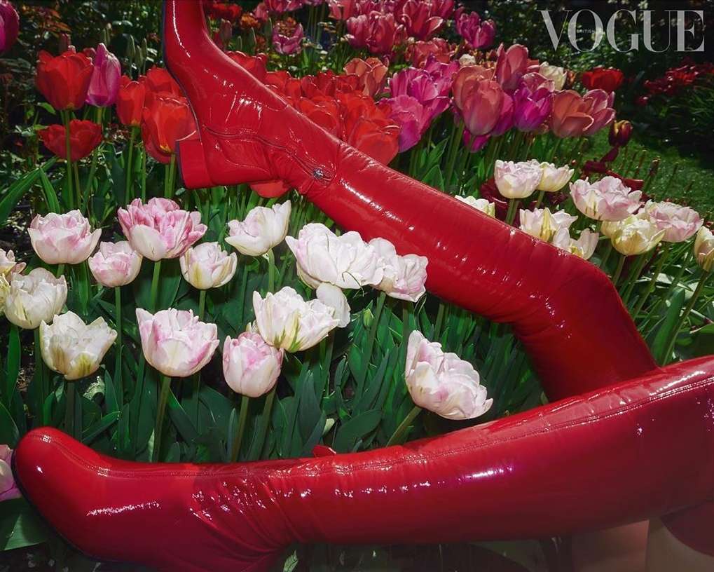 A pair of red, patent-leather boots are positioned in a field of red, pink, and white tulips with green stems and leaves, Vogue logo visible.