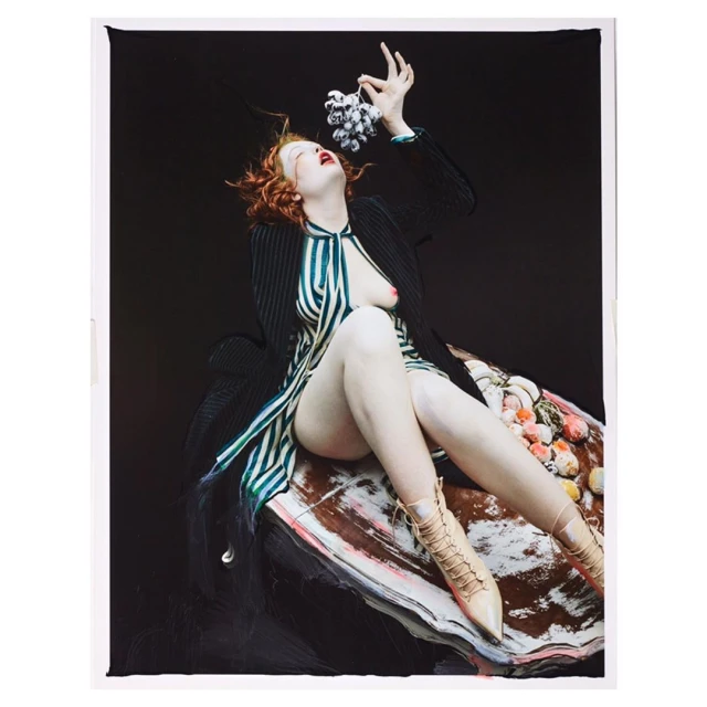 Woman with red hair sits partially nude on shell-like object holding grapes in a fine art, studio portrait against black backdrop.