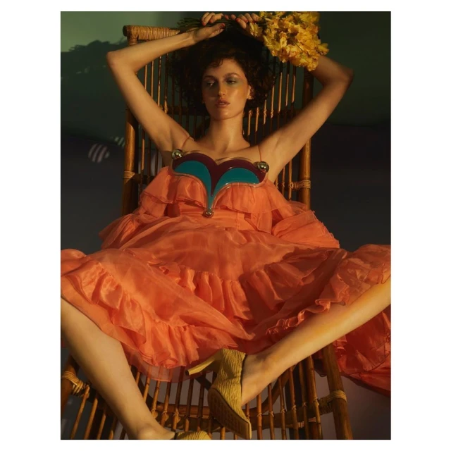A brunette woman sits in a rattan chair with a bunch of yellow flowers above her head. She is wearing an orange dress and yellow high heels.