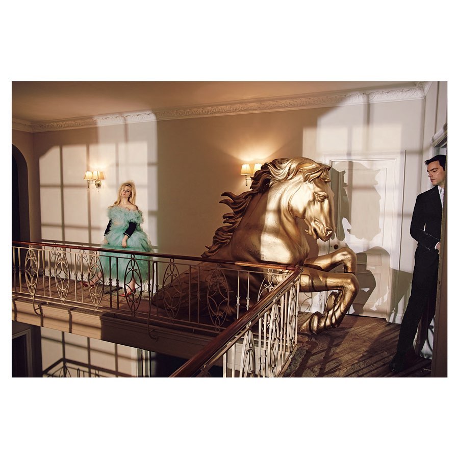 Indoor shot features a horse sculpture, an adult woman wearing a light blue dress, and an adult man in a black suit, all in a stylish setting.