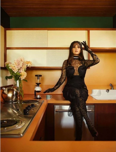Portrait of a woman in a black lace dress sitting on a kitchen counter with flowers, appliances, and orange cabinetry.