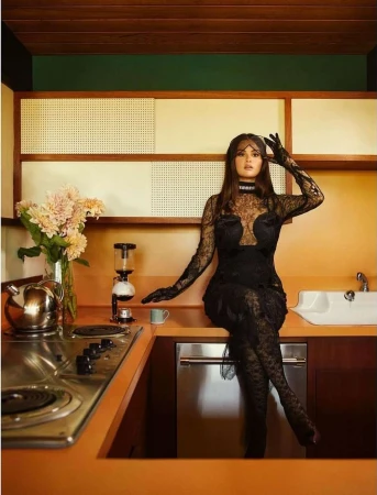 Portrait of a woman in a black lace dress sitting on a kitchen counter with flowers, appliances, and orange cabinetry.