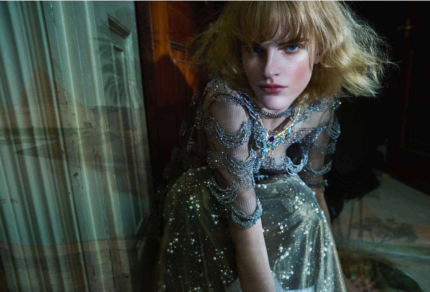 Indoor portrait of an adult woman with blond hair in a sparkly gown, posing expressively with direct eye contact in subdued lighting.