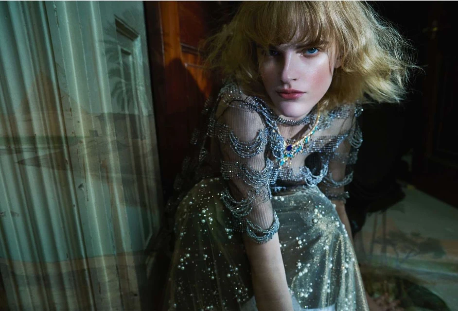 Indoor portrait of an adult woman with blond hair in a sparkly gown, posing expressively with direct eye contact in subdued lighting.