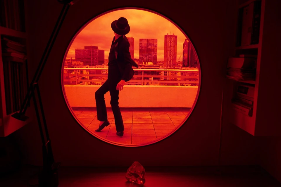 A stylish adult in a suit and hat poses on a balcony with city buildings in the background, framed by a circular window in a red-toned setting.