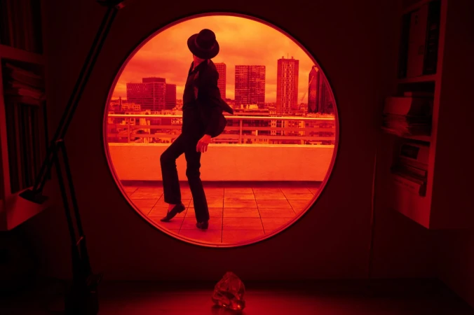 A stylish adult in a suit and hat poses on a balcony with city buildings in the background, framed by a circular window in a red-toned setting.