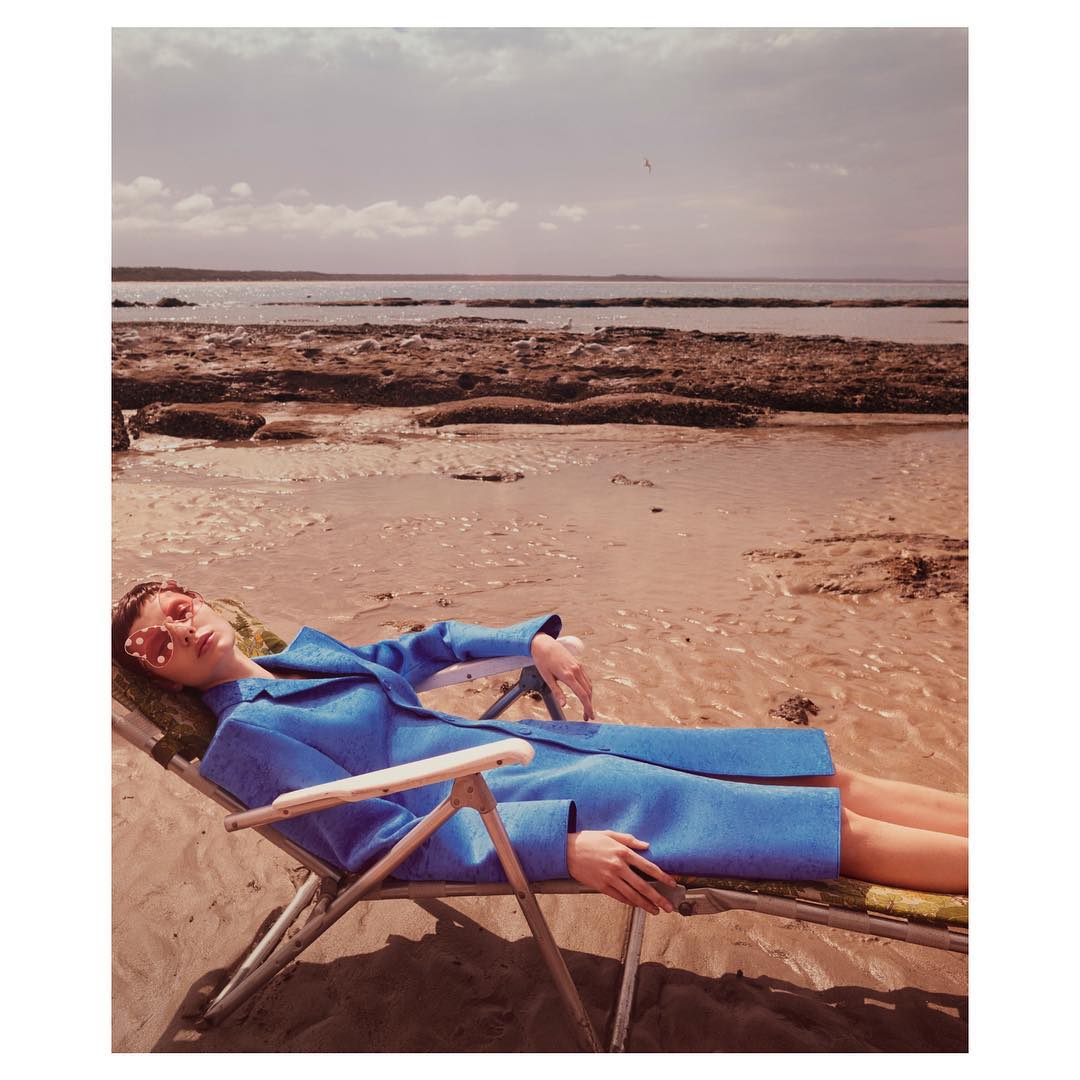 A woman in a blue coat lies on a beach chair on a sand beach by a rocky coastline under an overcast sky in a fashion editorial shot.