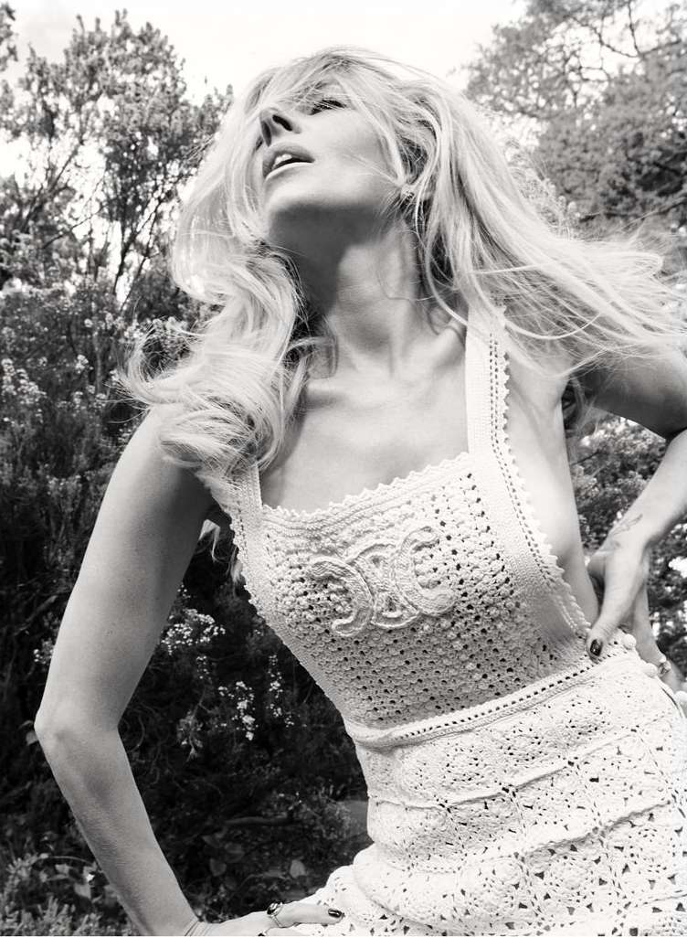 Monochrome portrait of a blonde adult woman with long wavy hair, wearing a crocheted dress, posing outdoors in a garden setting.